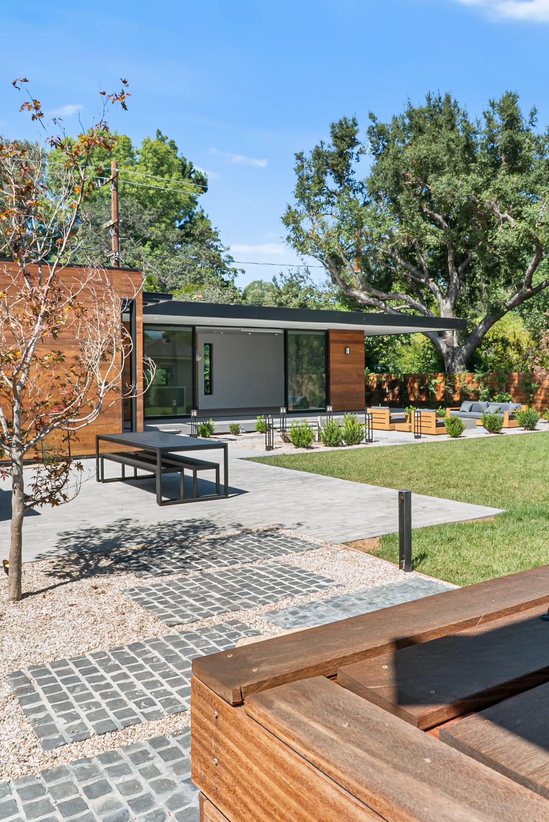 Modern single-story home with wood cladding and floor-to-ceiling glass walls, opening onto a paved patio and landscaped garden.