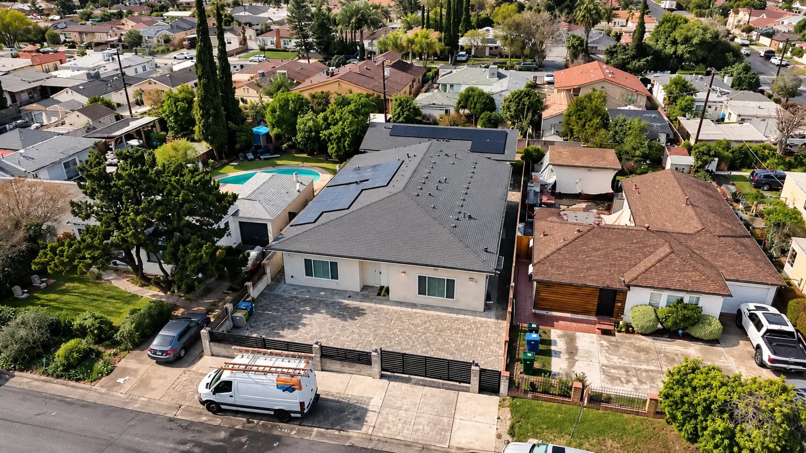 Aerial view of a residential property with rooftop solar panels, paved driveway, and surrounding suburban homes.